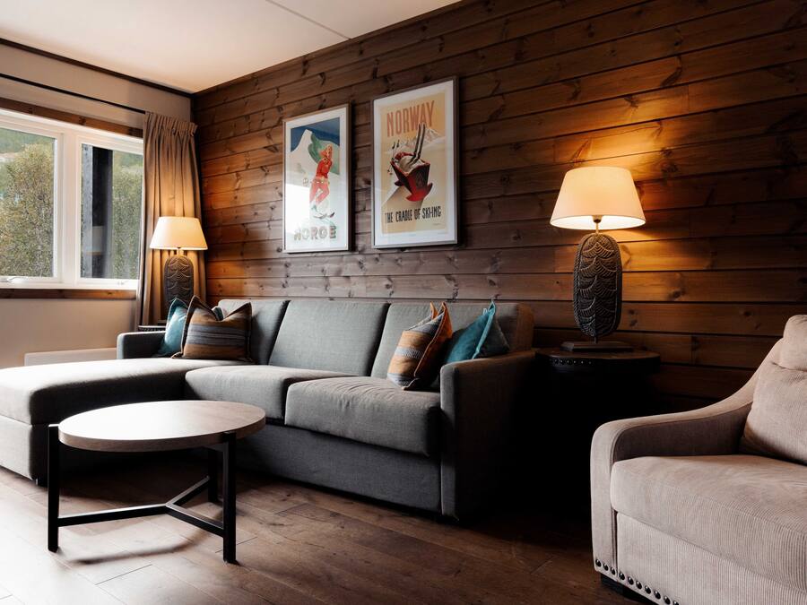 Ein modernes Wohnzimmer mit einem grauen Sofa, einem runden Couchtisch und zwei Beistelltischen mit Lampen. An der Wand hängen zwei gerahmte Poster, eines davon trägt die Aufschrift "Norway".