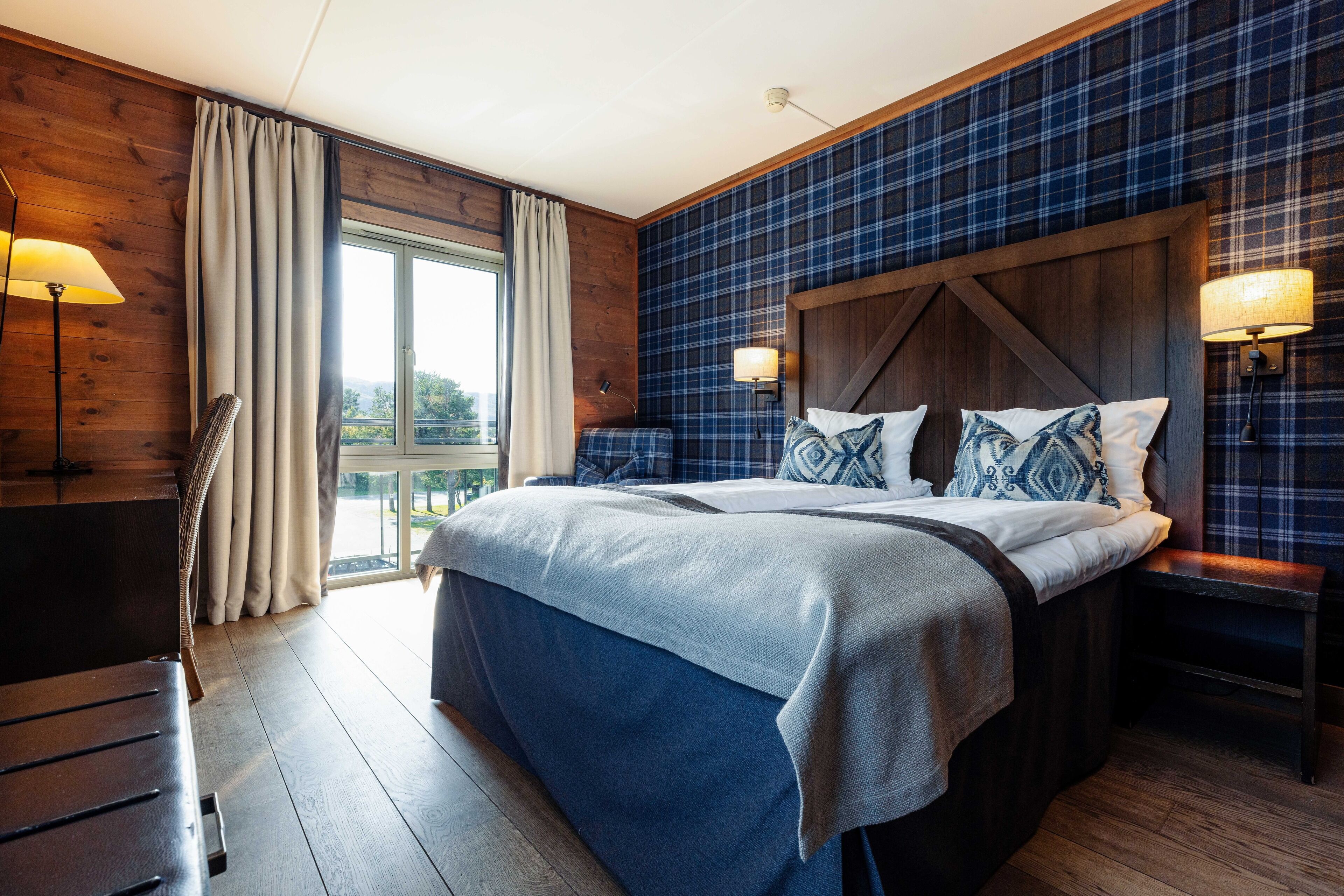 A bedroom with a large bed, wooden headboard, plaid wallpaper, and a view through the window.