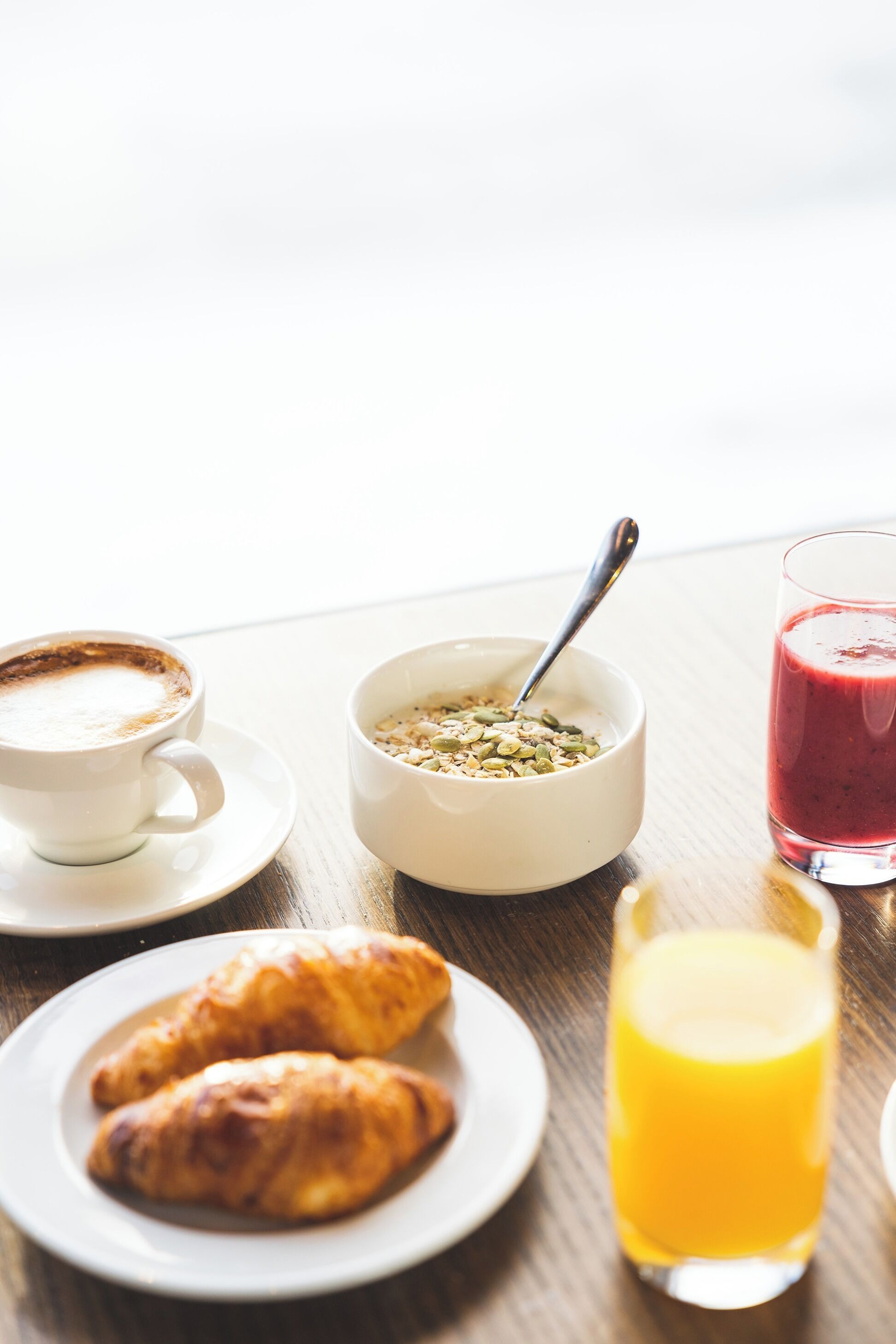 Une table en bois dressée pour le petit-déjeuner, avec une tasse de café, un bol de céréales, un verre de jus d’orange et deux croissants.