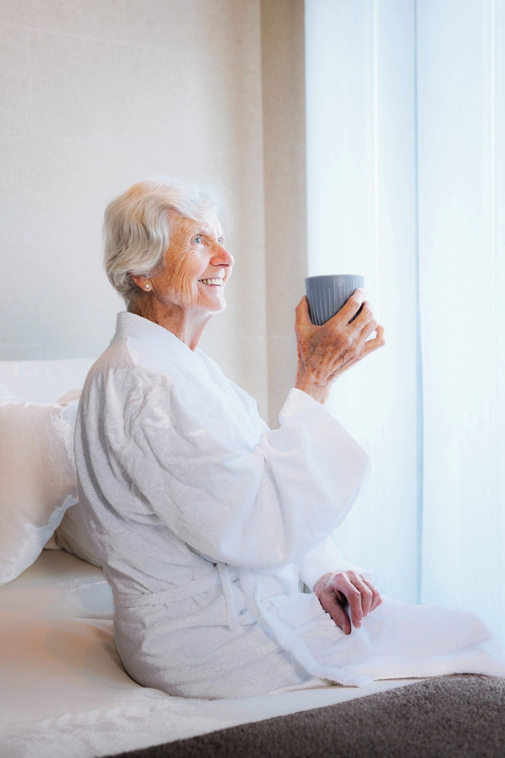 Una persona mayor sentada en una cama con una bata blanca, sosteniendo una taza.