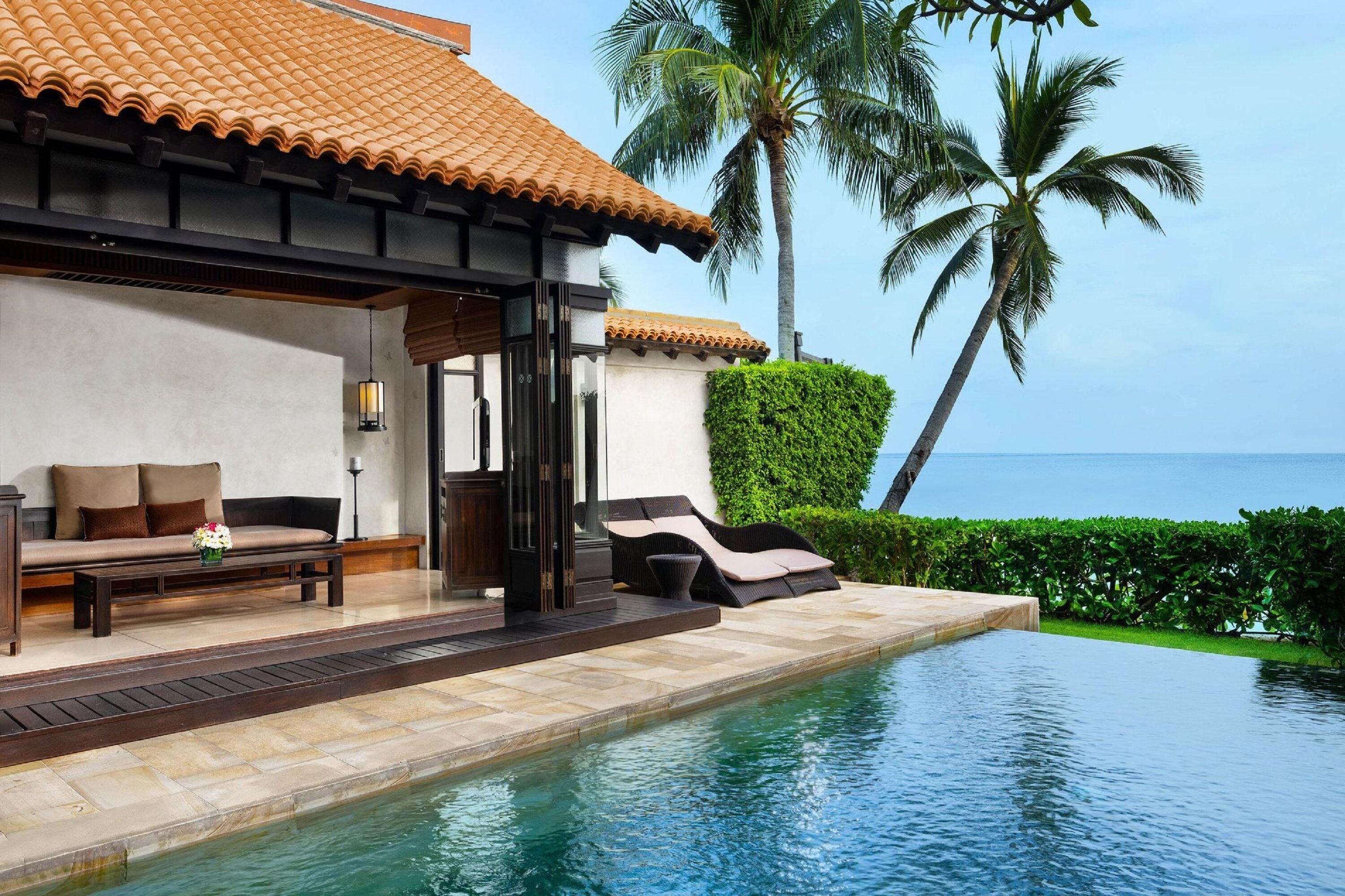 A poolside area with a sofa and lounge chairs, a tiled roof, and a view of the ocean.