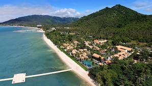 An aerial view of a coastal resort with a beach, pool, and lush greenery.