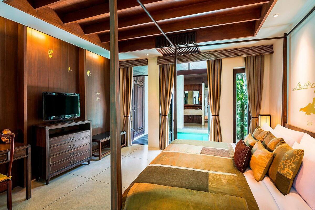 A modern hotel room with a wooden ceiling, a large bed with multiple pillows, a TV on a wooden cabinet, and a view of a pool and greenery through the curtains.