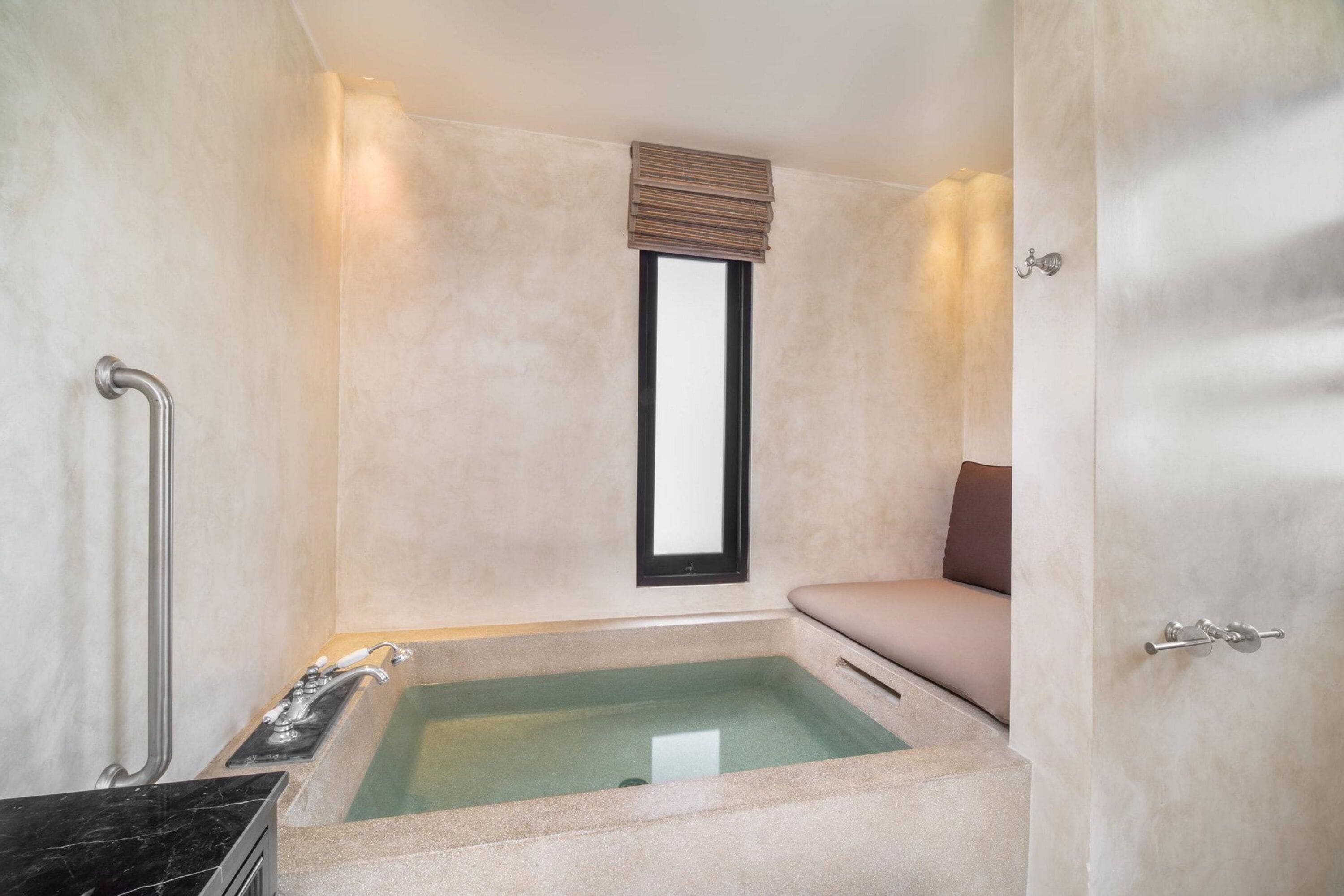 Une salle de bain avec une baignoire, une fenêtre avec des stores, un porte-serviettes et une porte avec une poignée.