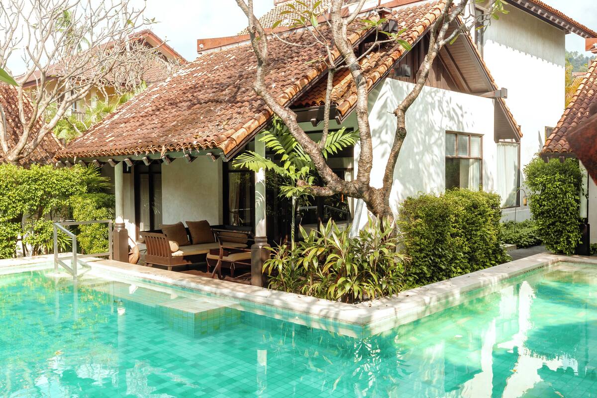 A swimming pool with tiled edges, surrounded by greenery and a building with a tiled roof.
