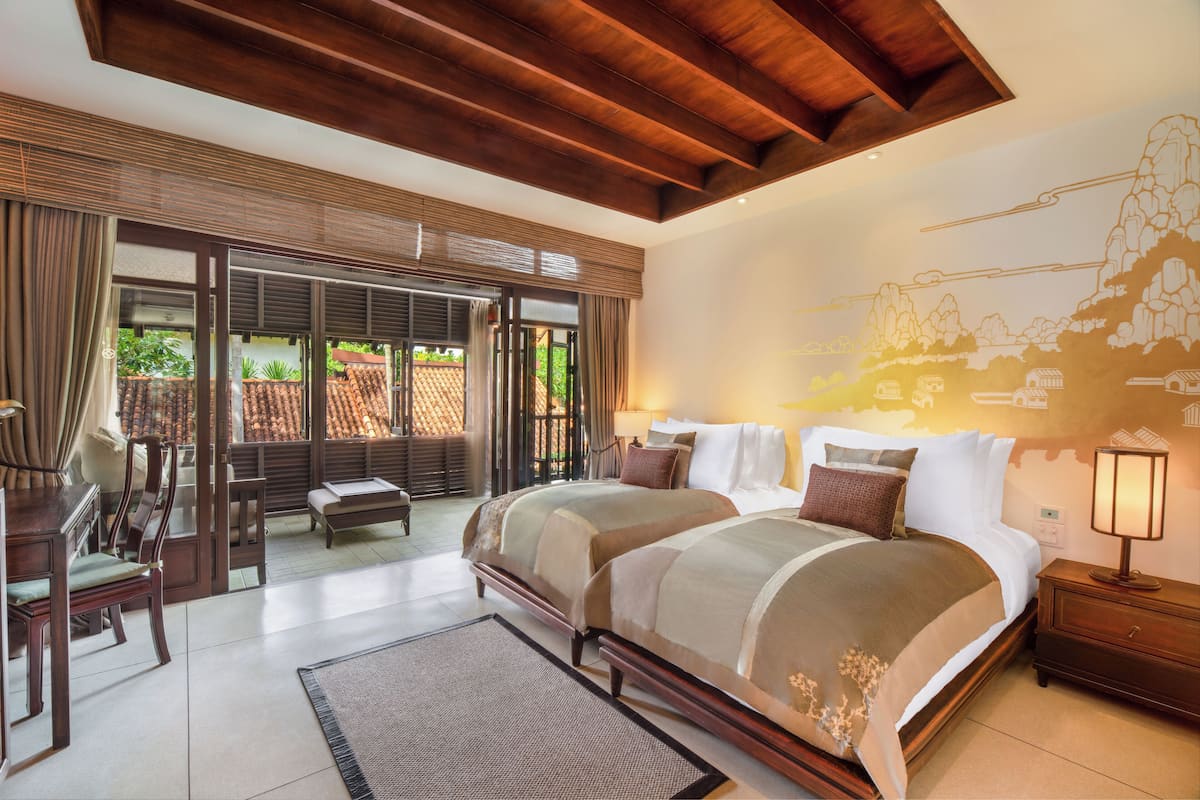 A bedroom with a large bed, a desk, and a chair. There is a window with curtains and a view of greenery outside.