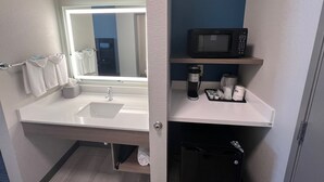 A modern bathroom with a white sink, a mirror with a light, a black trash bin, and a shelf with a microwave and toiletries. - Holiday Inn Express Wichita North - Park City by IHG (Wichita)