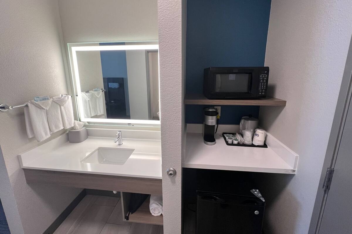 Une salle de bain moderne avec un lavabo blanc, un miroir avec éclairage intégré, une poubelle noire et une étagère sur laquelle se trouvent un micro-ondes et des articles de toilette.