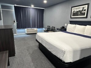 A hotel room with a bed, a desk, a chair, a desk lamp, a telephone, and a window with curtains.