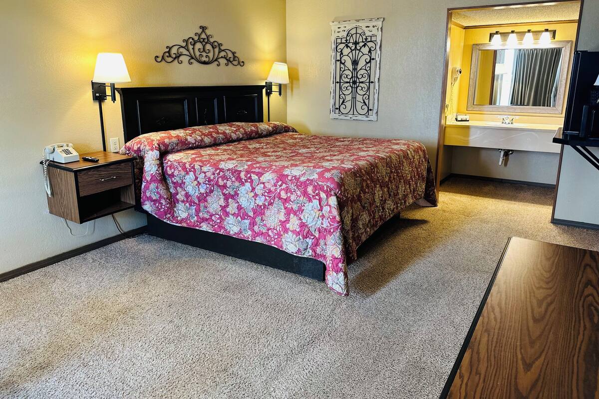 A hotel room with a bed, bedside lamps, a telephone, a mirror with lights, and a decorative wall piece.
