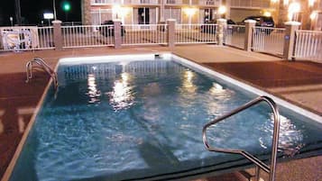 Una piscina rectangular con barandillas metálicas, rodeada por una valla blanca. Al fondo, se observa un edificio con varios balcones y luces encendidas.