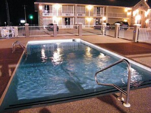 A rectangular swimming pool with metal railings, surrounded by a white fence. In the background, there is a building with multiple balconies and lights on.