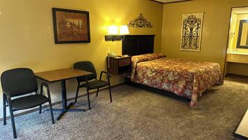 A hotel room with a bed, a desk, two chairs, a lamp, a telephone, and a picture on the wall.
