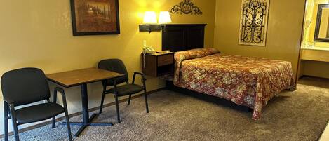 A hotel room with a bed, a desk, two chairs, a lamp, a telephone, and a picture on the wall.