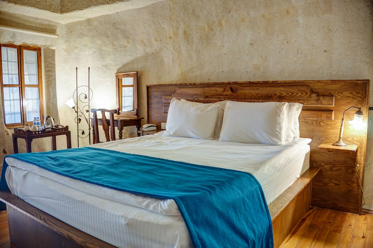 A bedroom with a wooden headboard, a bed with white linens and a blue blanket, a bedside table with a lamp, and a small wooden nightstand.