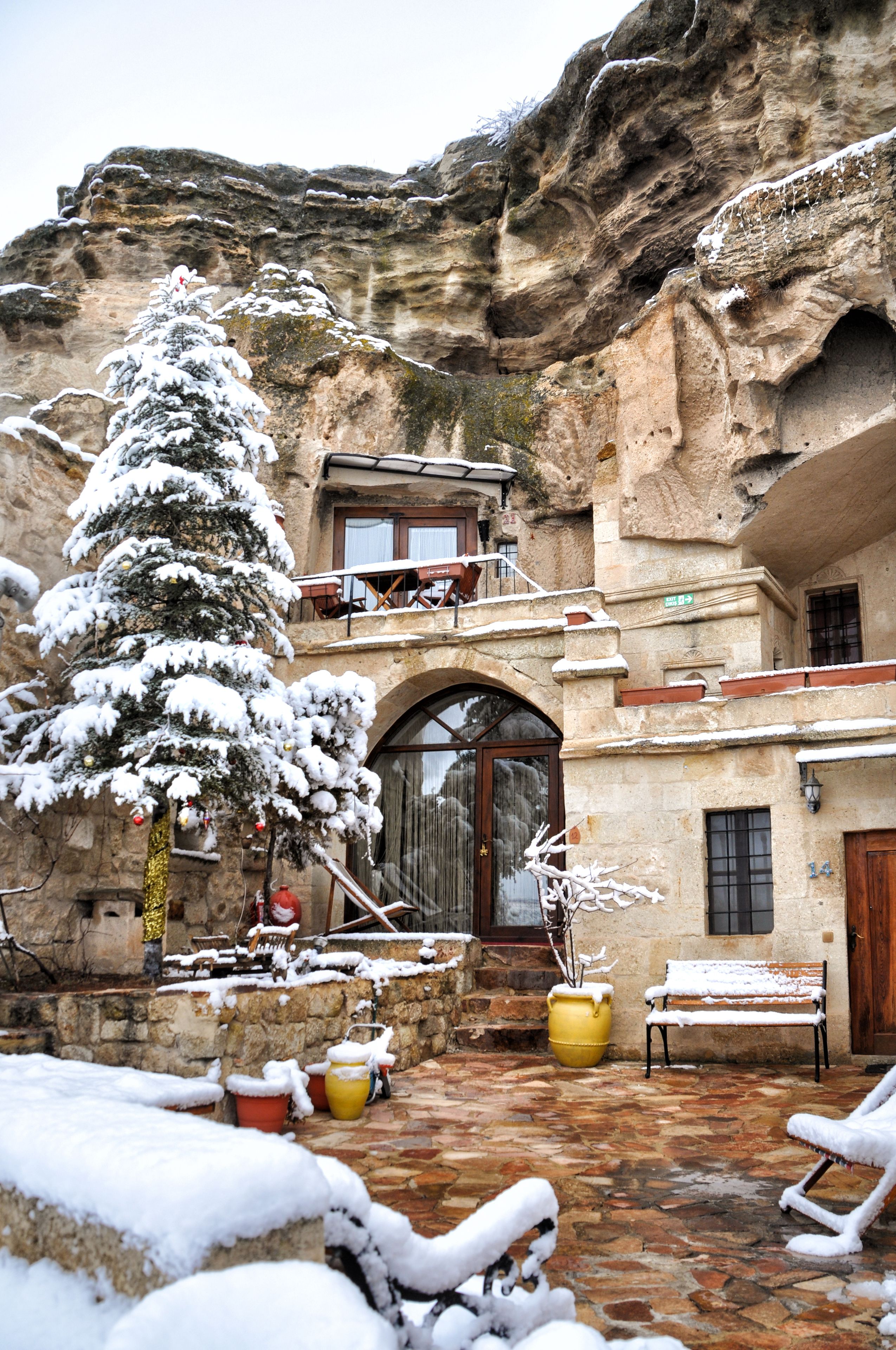 Une cour enneigée avec un bâtiment en pierre, un arbre recouvert de neige et des sièges installés à l’extérieur.
