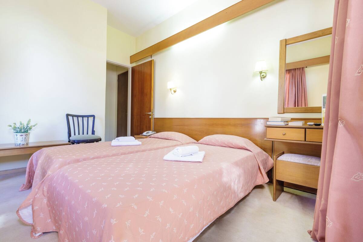 A hotel room with two beds, a wooden headboard, a bedside table, a chair, a small table with a plant, and a mirror.
