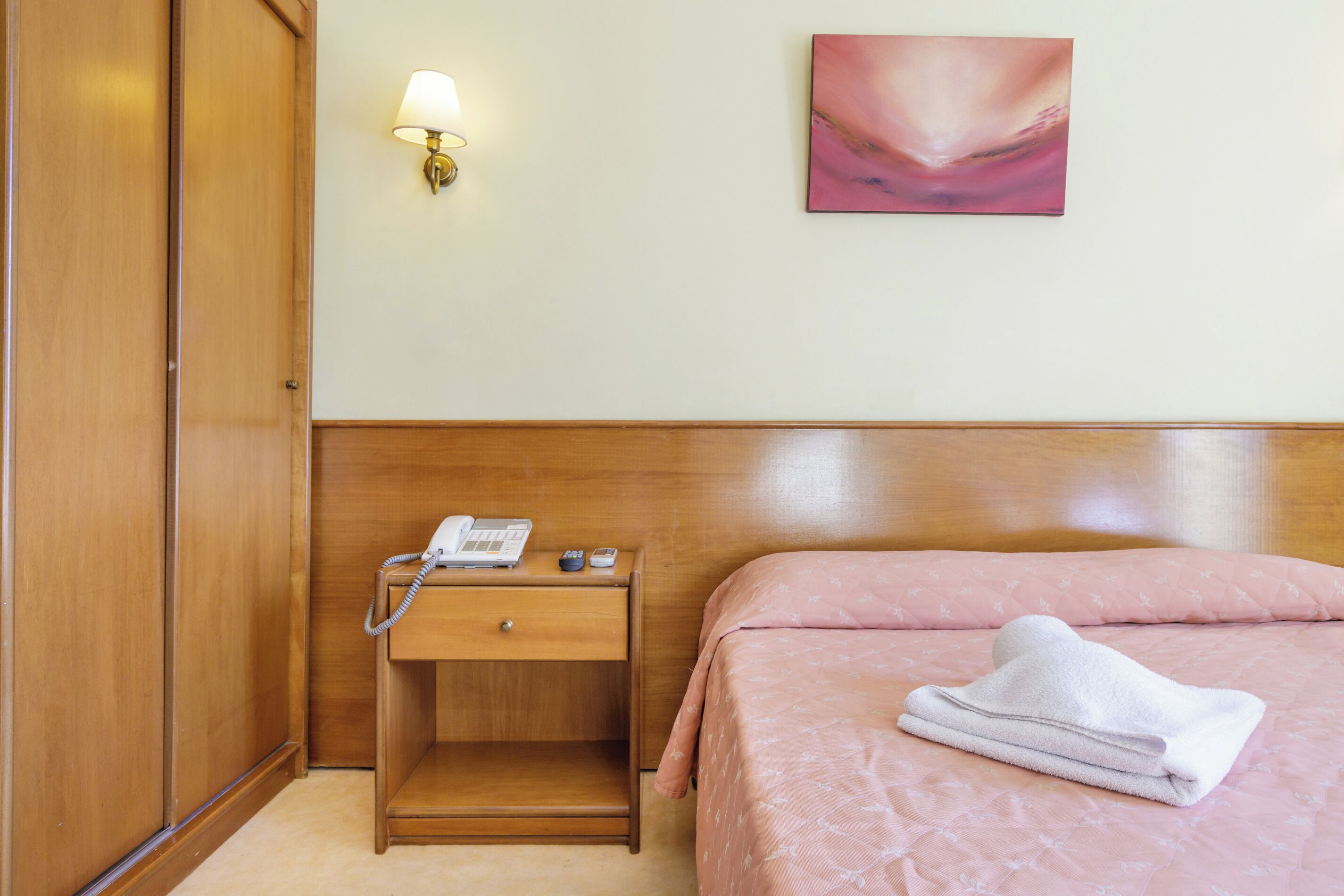 Une chambre d’hôtel avec une armoire en bois, une table de chevet avec une lampe, un téléphone et une serviette pliée sur le lit.