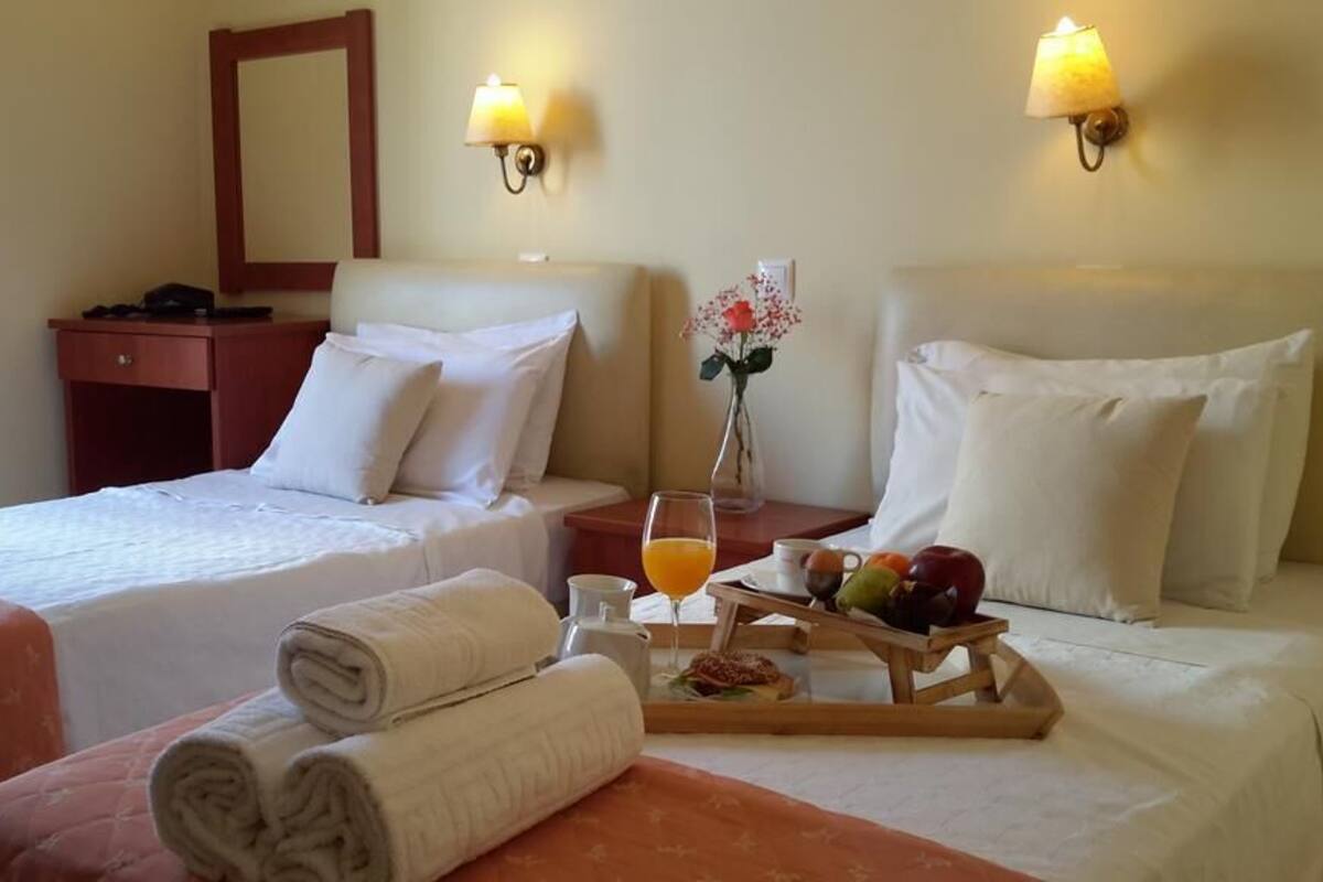 A hotel room with two beds, a bedside table with a lamp, a tray with breakfast items, and a vase with flowers.