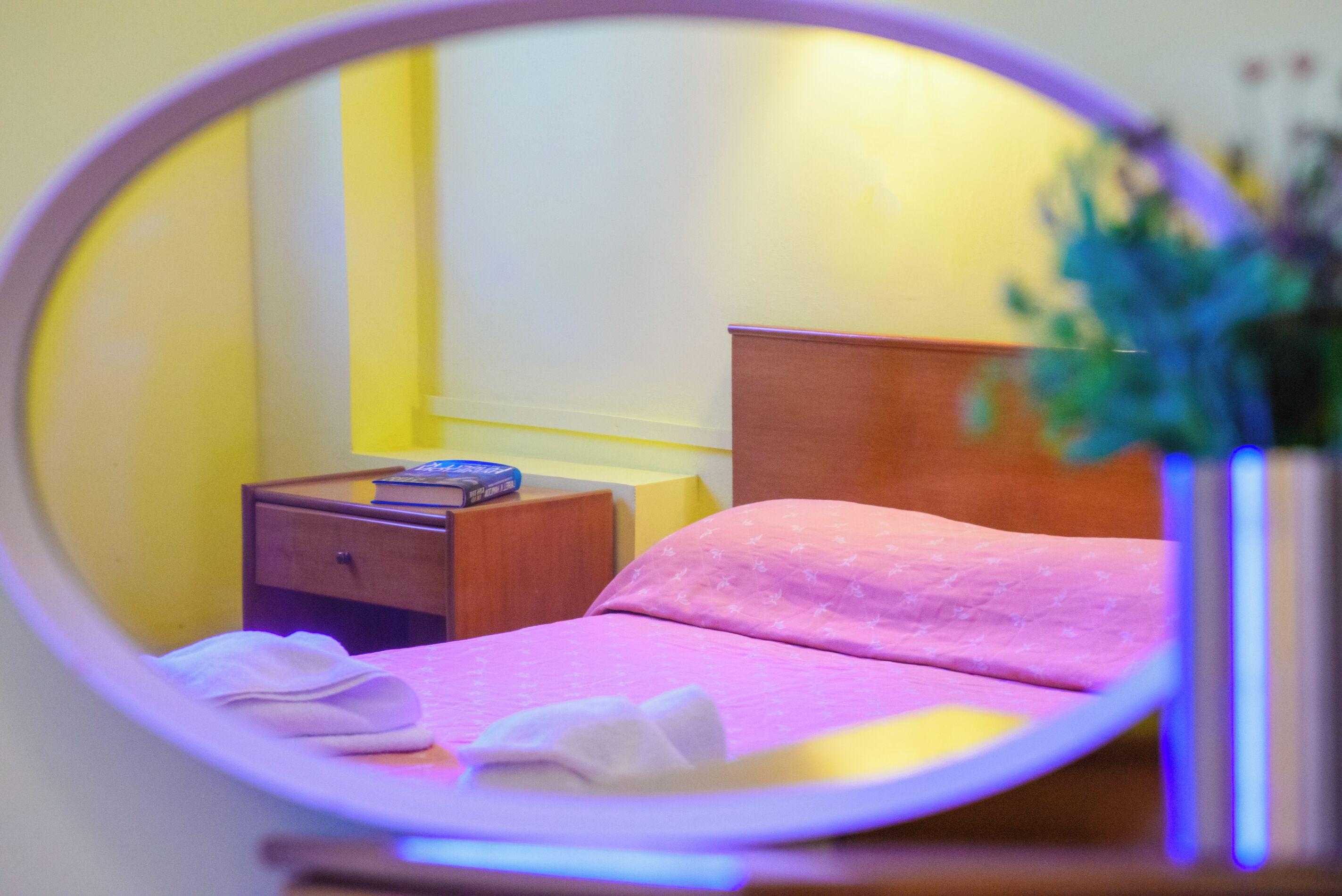 Um espelho circular reflete uma cama com colcha rosa, uma mesa de cabeceira de madeira com um livro e uma pequena planta.