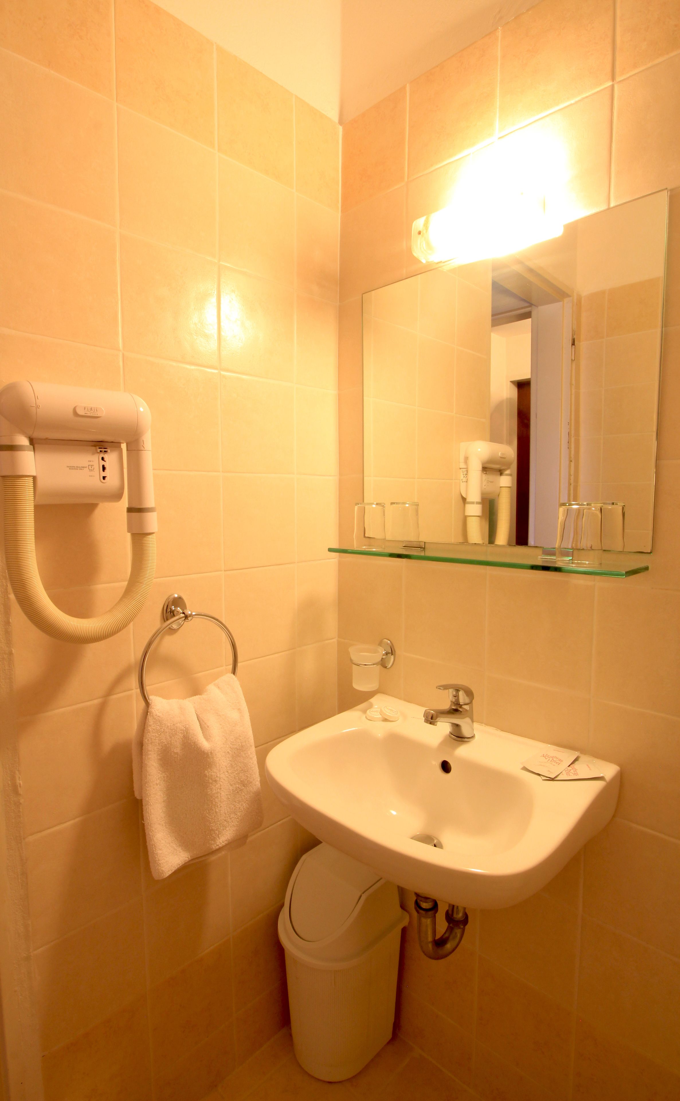 Un baño con un lavabo blanco, un estante de vidrio encima, un perchero y un secador de pelo en la pared.