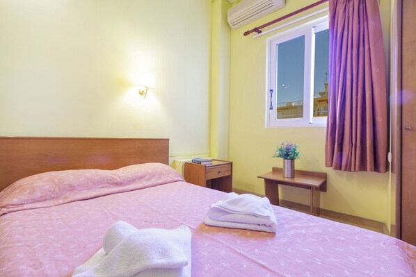 Una cama bien hecha con ropa de cama rosa y una toalla doblada, una mesita de noche con un libro y un florero, y una ventana con cortinas moradas.