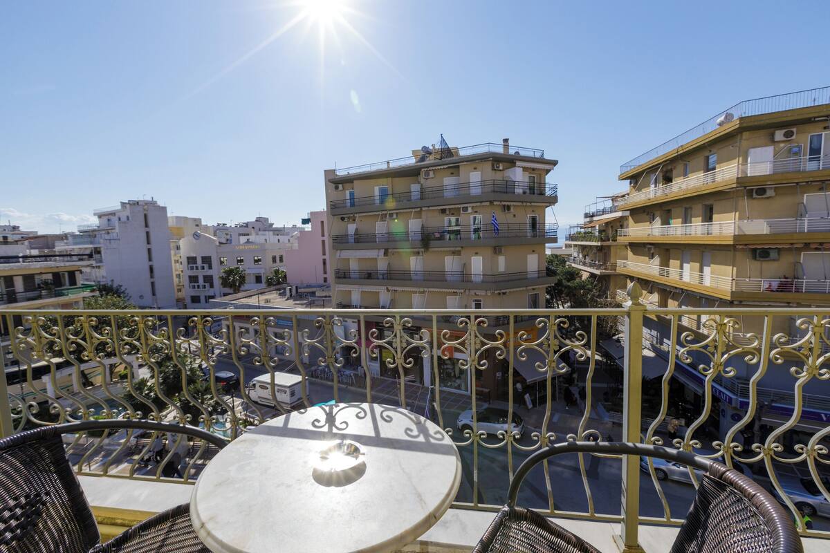 A balcony with a table and chairs, overlooking a street with buildings and parked cars.