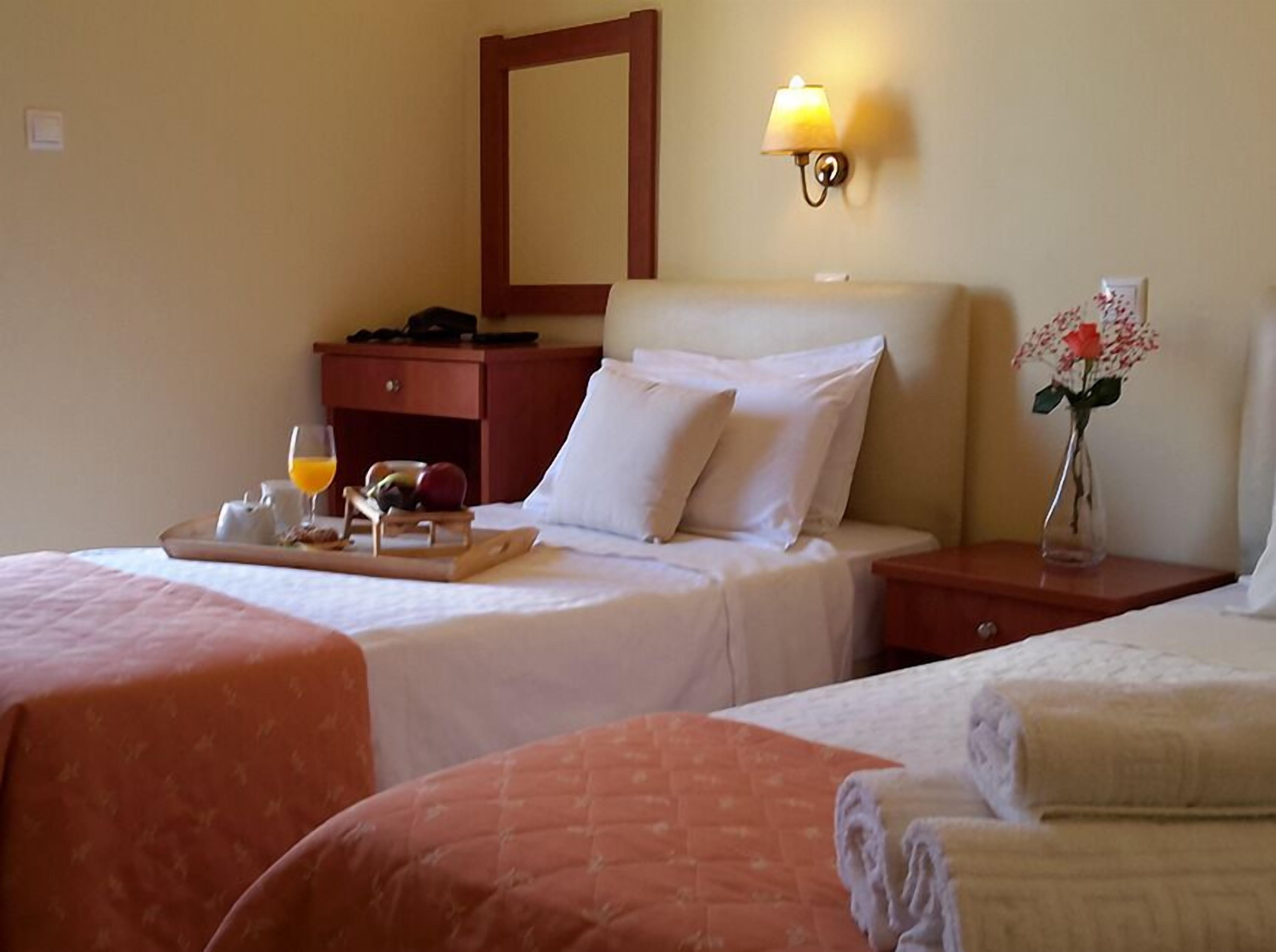 A hotel room with two beds, a bedside table with a lamp, a tray with breakfast items, and a vase with flowers.