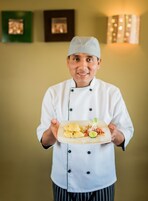 Un chef con uniforme blanco sostiene un plato rectangular con comida.