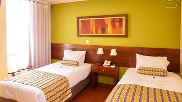 Habitación de hotel con dos camas, cada una con ropa de cama estampada, cabeceros de madera, mesitas de noche con teléfono y un cuadro en la pared.