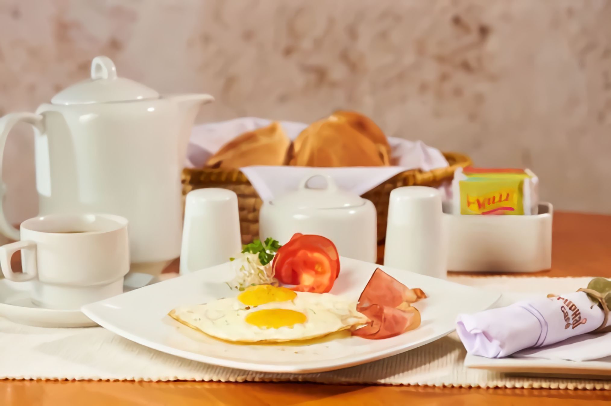 A breakfast setting with a plate of eggs, tomato, and ham, a teapot, cups, and a basket of bread.