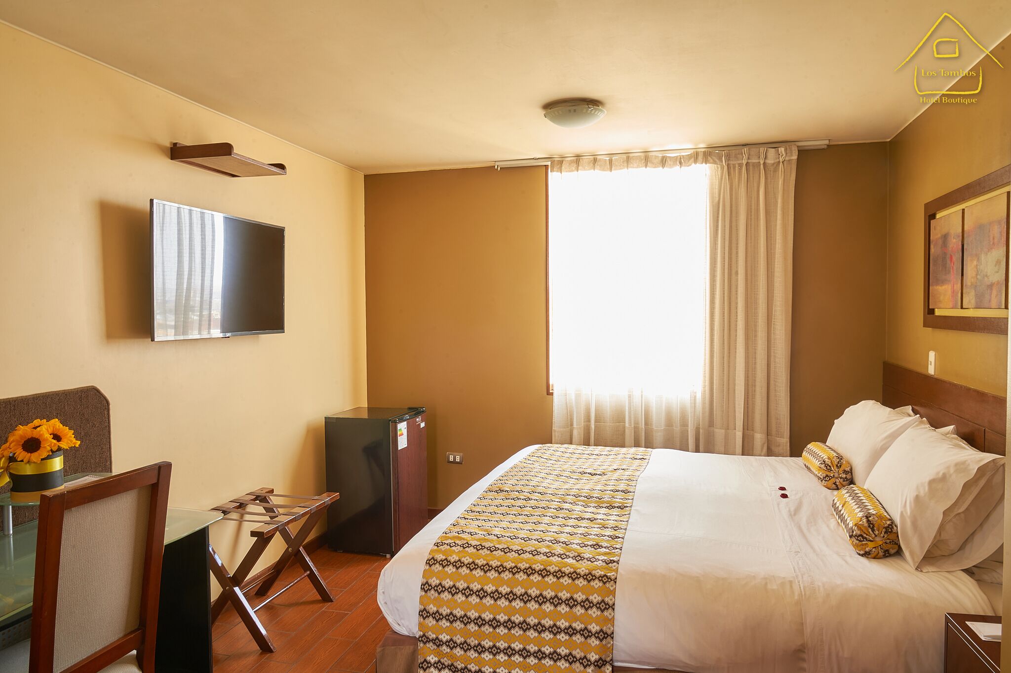 A hotel room with a bed, a TV mounted on the wall, a small table with a vase of sunflowers, and a window with curtains.