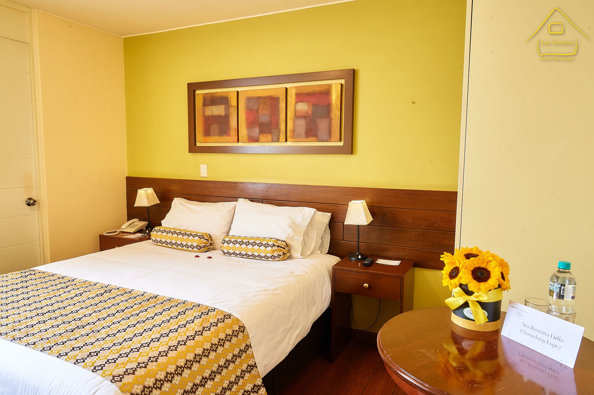 Habitación de hotel con cama, mesita de noche, lámpara, un jarrón con girasoles y una botella de agua.
