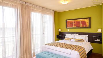 Una habitación de hotel con cama, mesita de noche, lámpara, un cuadro enmarcado y una ventana con cortinas traslúcidas.