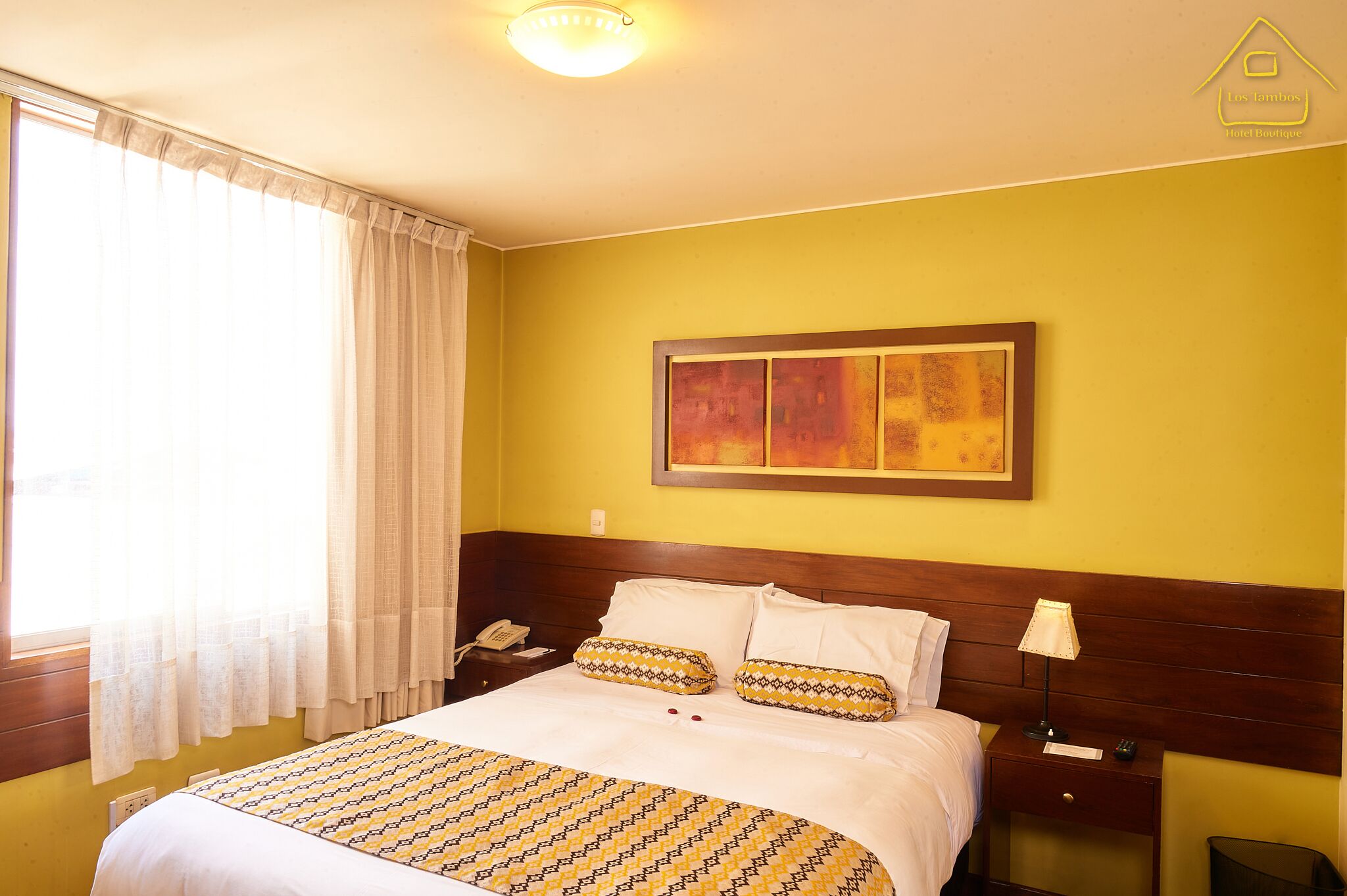 Habitación de hotel con cama, mesita de noche, lámpara, un cuadro en la pared y ventana con cortinas.