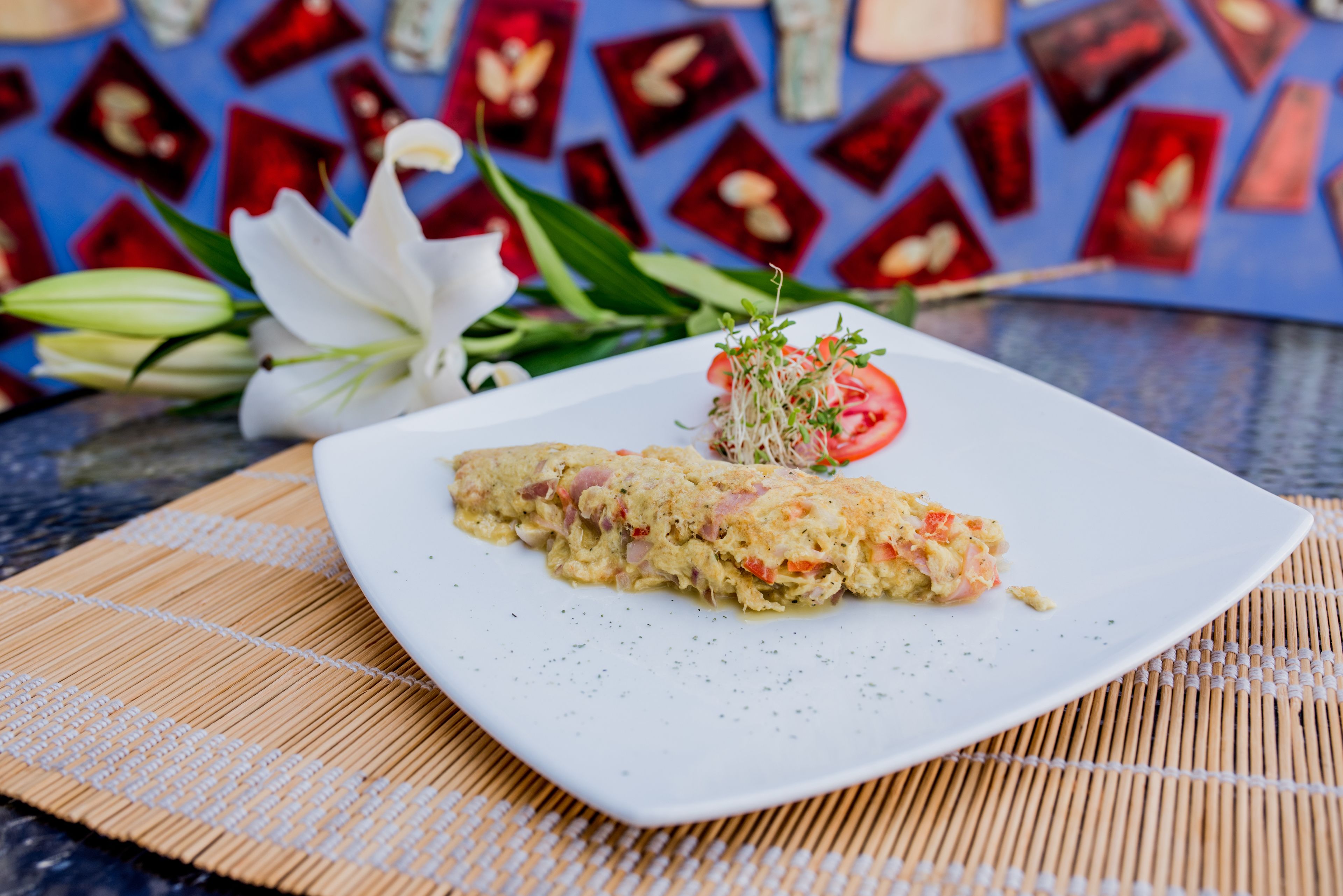 A square white plate with a slice of food, possibly a seafood omelette, on a bamboo mat.