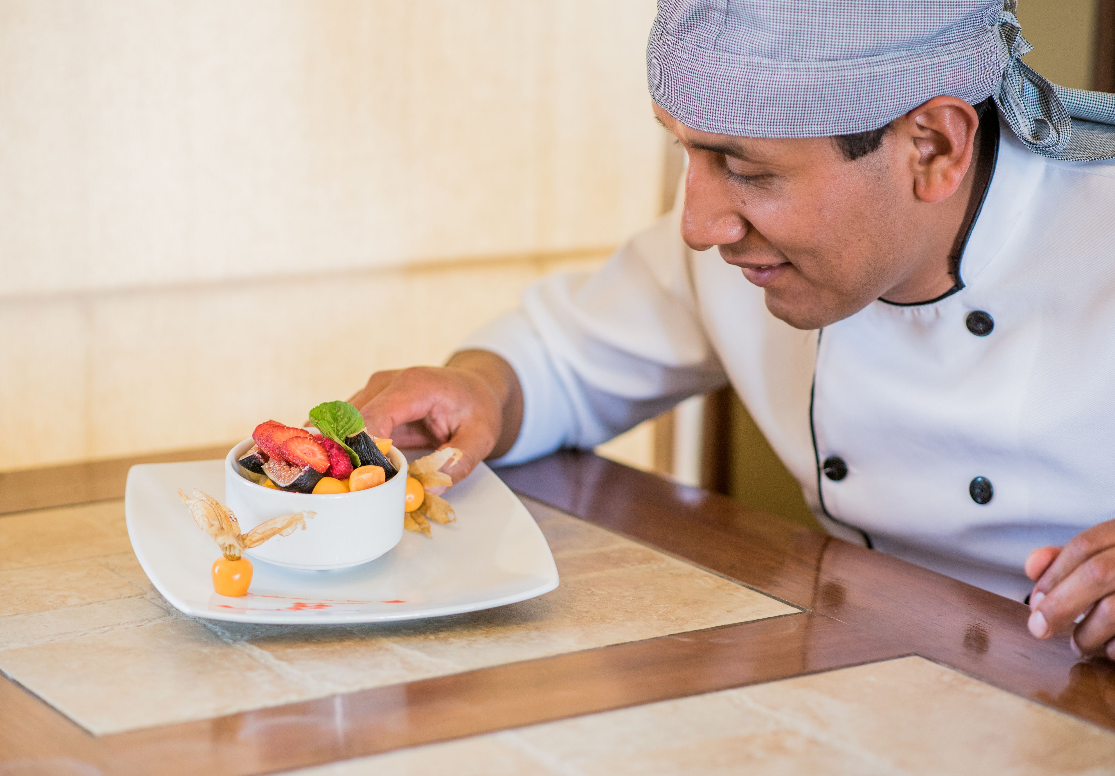 A chef in a white uniform with a checkered headband, preparing a dish on a white plate with a garnish.