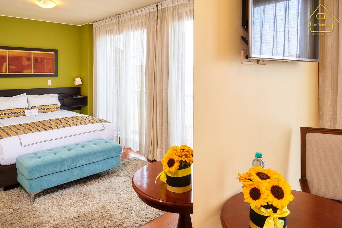 A hotel room with a bed, a blue bench, a table with a vase of sunflowers, and a TV on the wall.