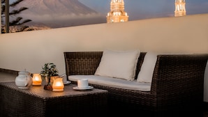 A wicker sofa with cushions, a small table with a teapot and candles, and a view of two lit towers against a mountain backdrop.