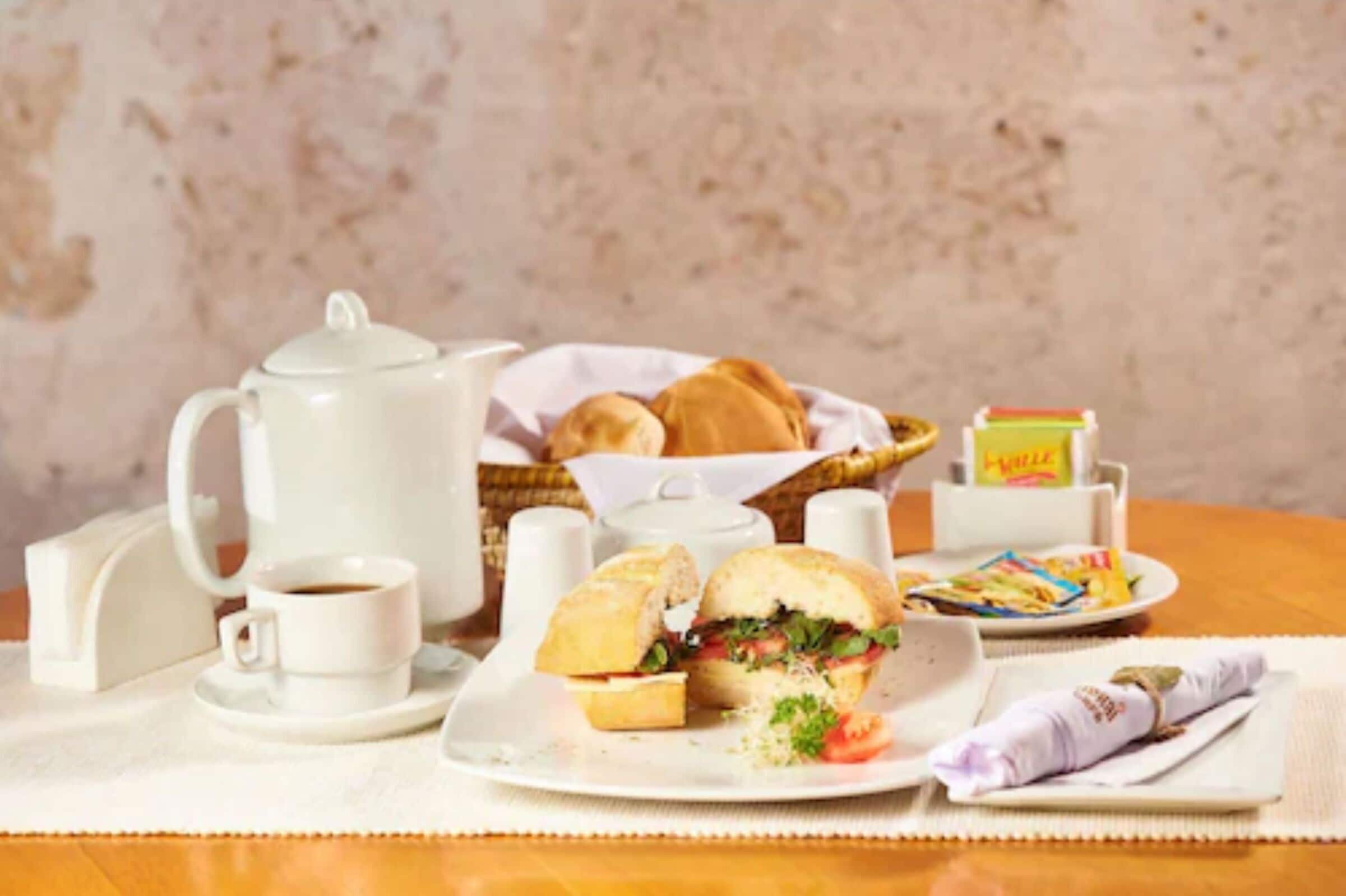 A breakfast setup with a teapot, cups, sandwiches, and a basket of bread.