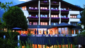 A multi-story hotel with balconies adorned with flowers, a covered outdoor seating area, and a view of mountains in the background.