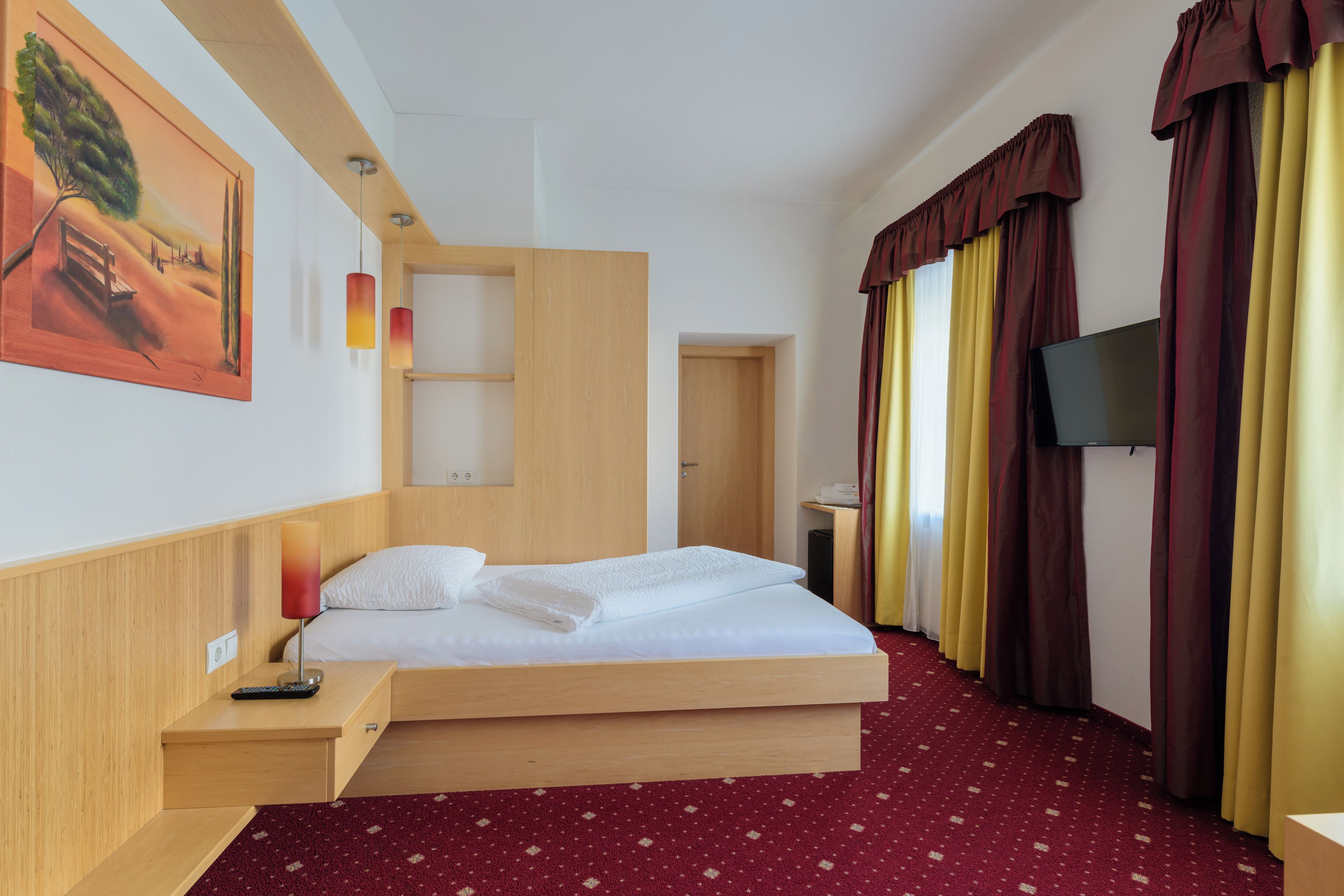 A hotel room with a bed, a desk, a TV, and a painting on the wall.