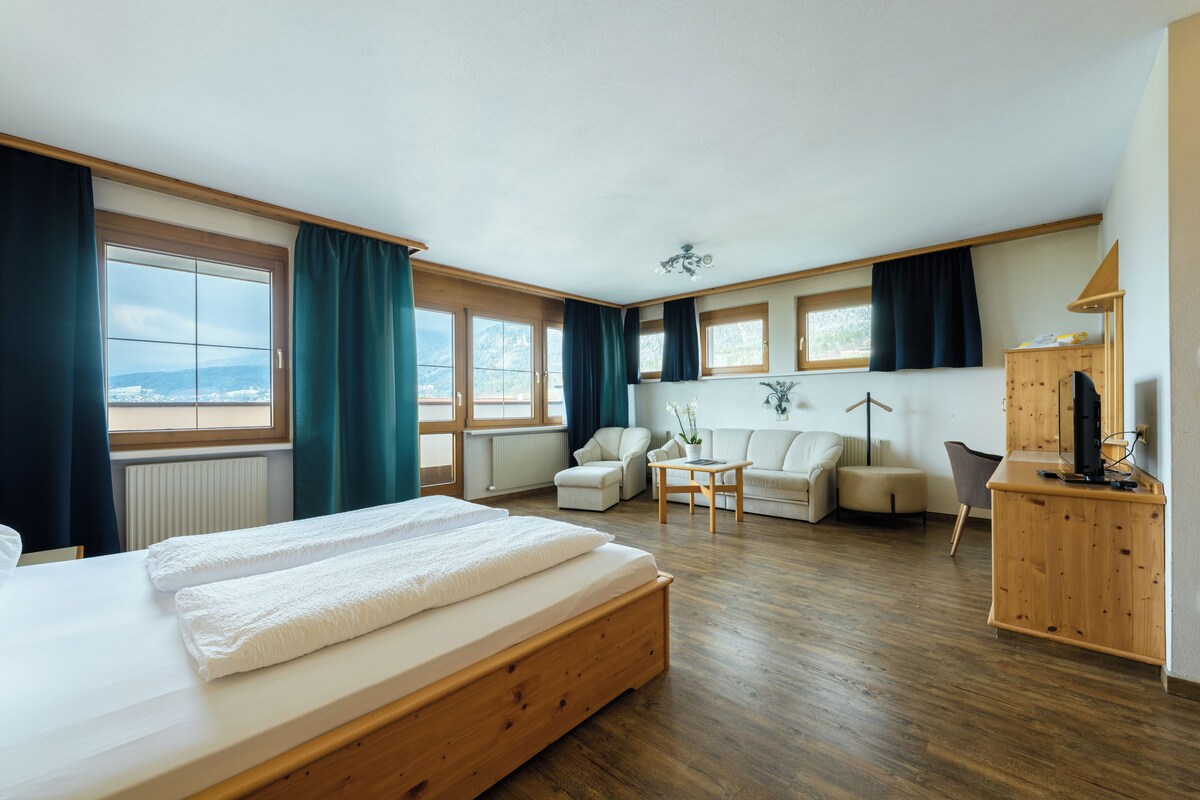 Un dormitorio amplio con una cama grande, un escritorio de madera, una silla y un sofá. Varias ventanas ofrecen luz natural y vistas al exterior.