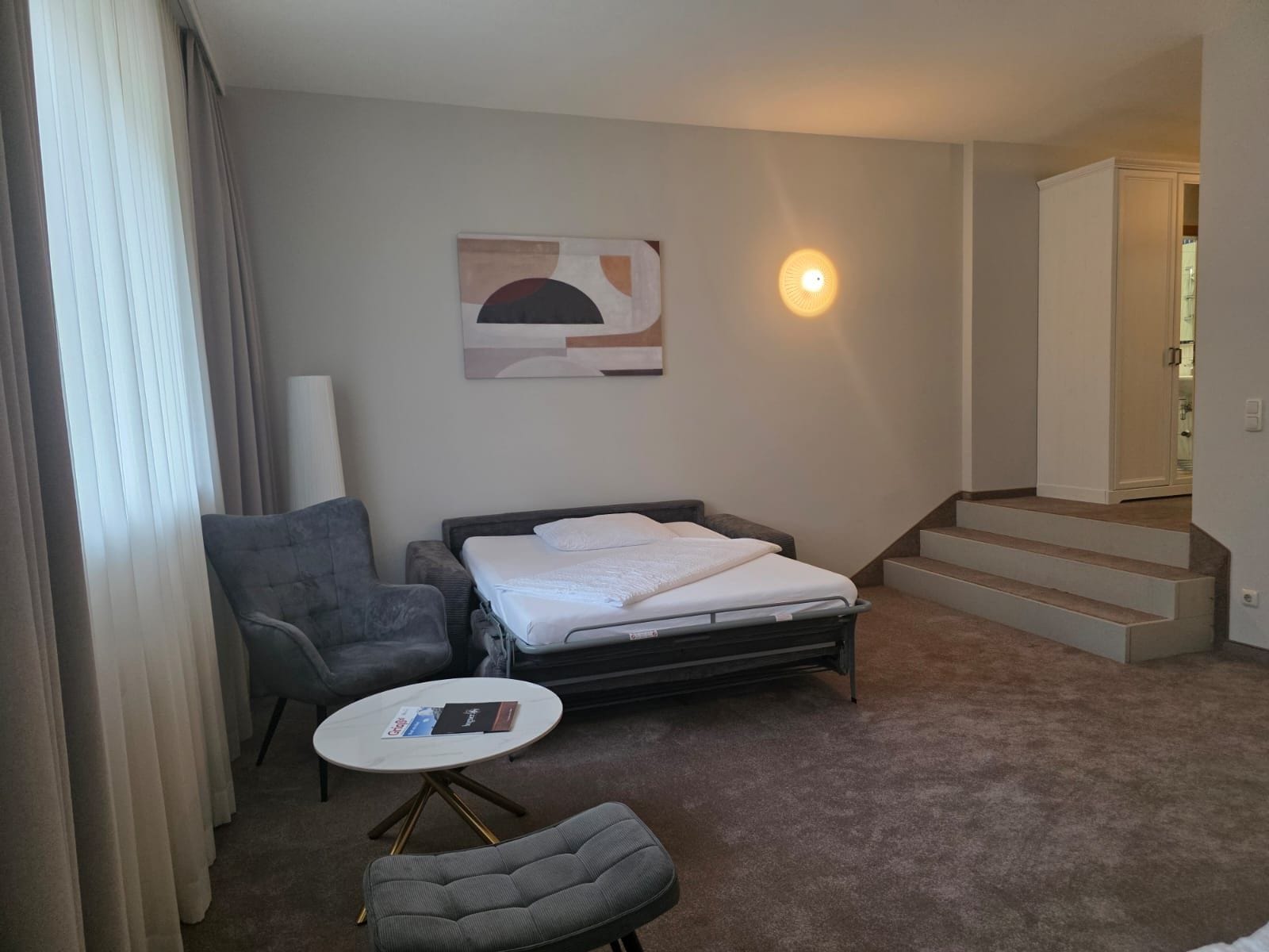 A hotel room with a bed, a chair, a small table, a painting on the wall, and a staircase leading to another room.