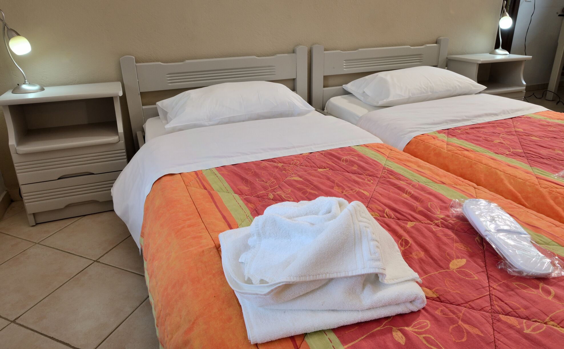 Cama doble con colcha roja y naranja, dos almohadas blancas, una toalla blanca doblada y una bolsa de plástico sobre la cama.