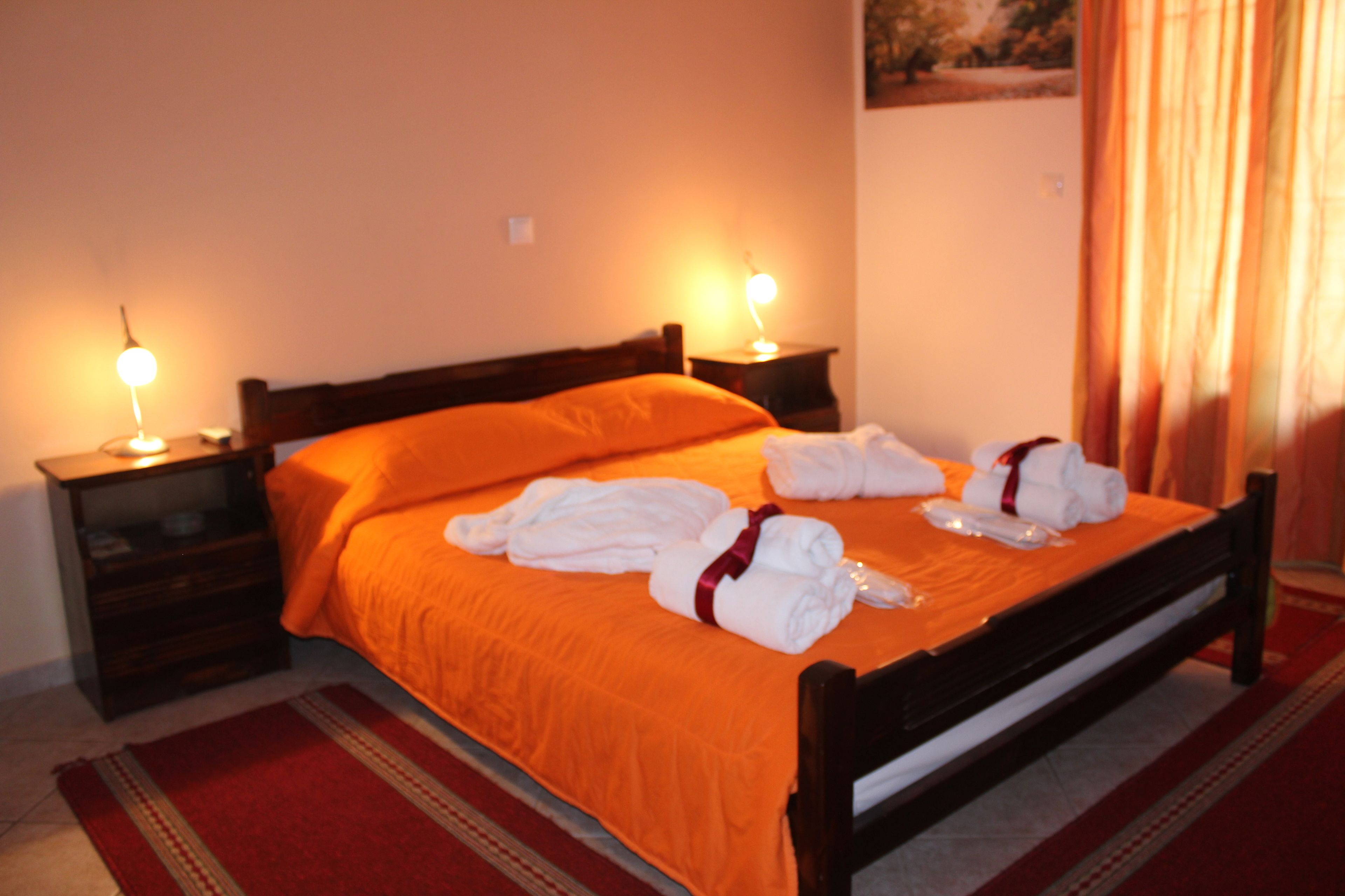 A neatly made bed with an orange bedspread and white towels, two bedside lamps, and a red carpet.
