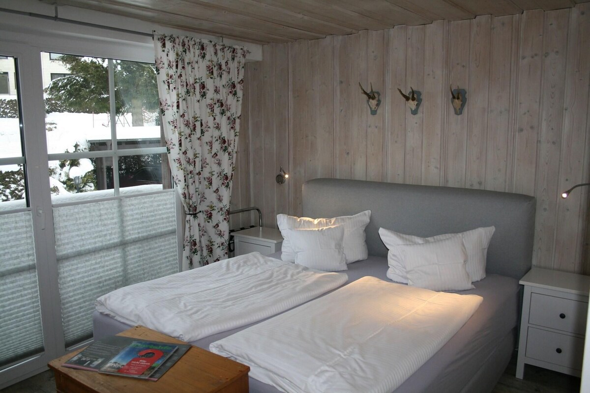 A bedroom with a bed, two bedside tables, a nightstand, a window with a curtain, and a view of the outside.