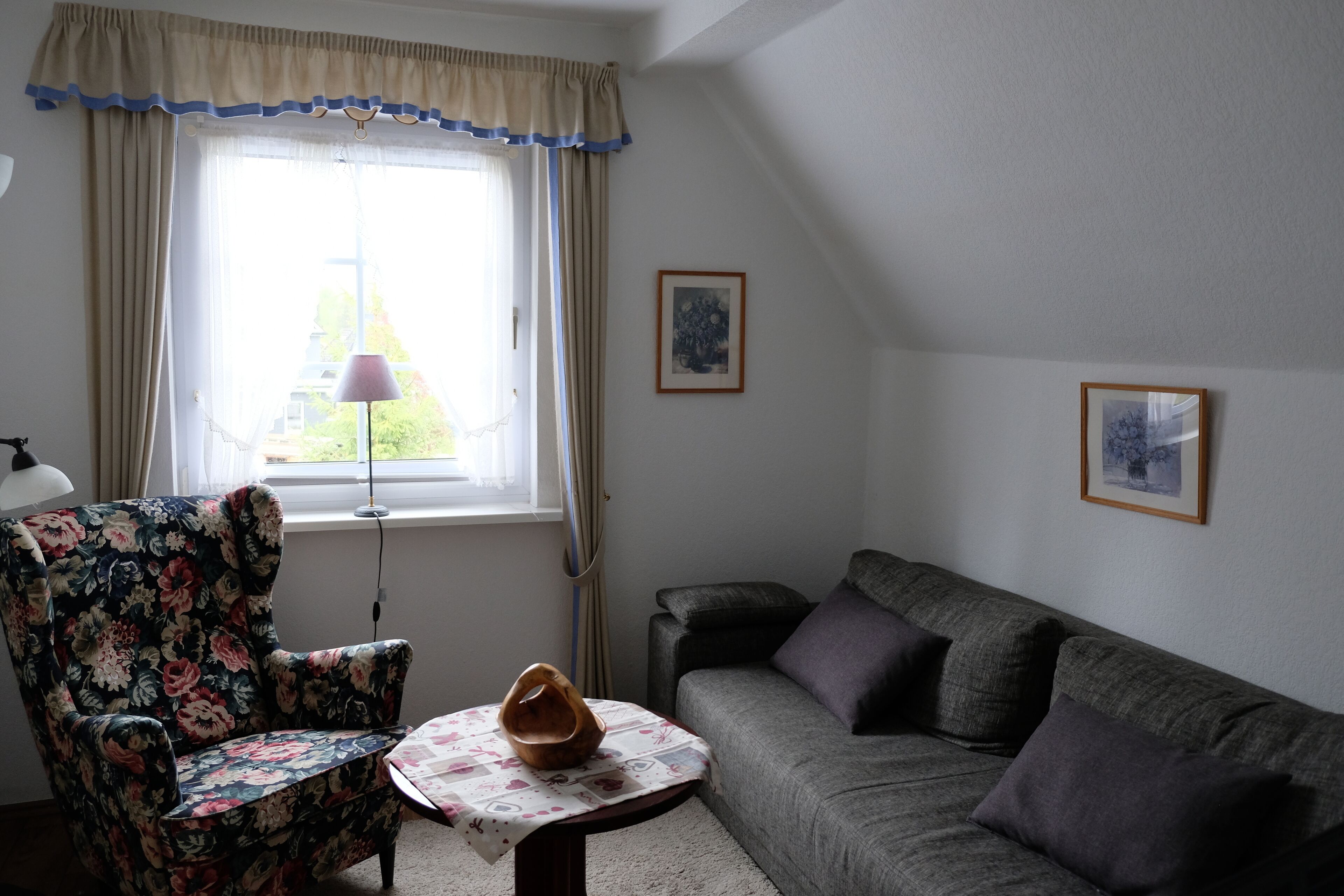 A living room with a floral armchair, a grey sofa, a small table with a vase, and two framed pictures on the wall.