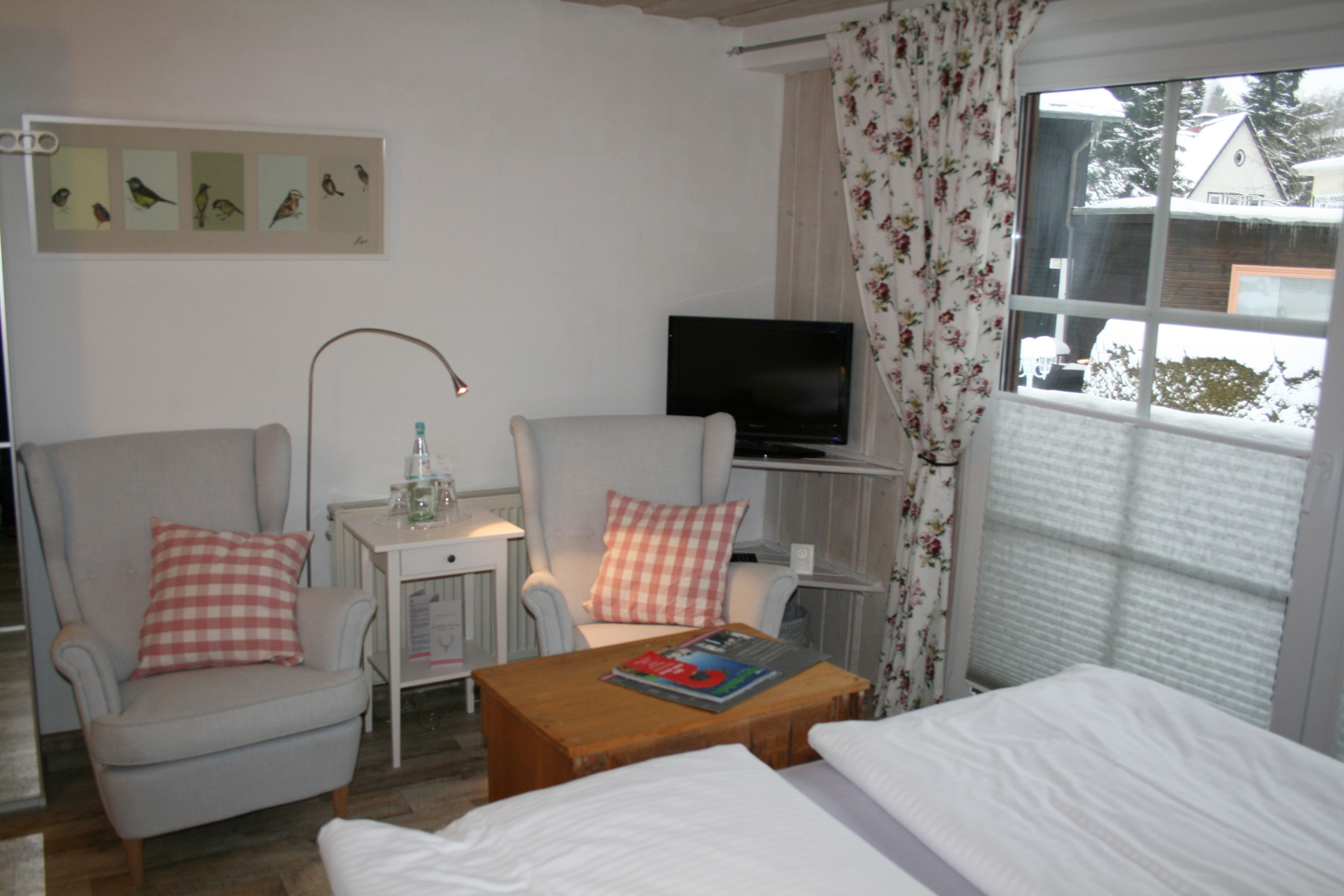 Une chambre d’hôtel avec deux fauteuils, une petite table sur laquelle repose un magazine, un téléviseur posé sur un meuble et une fenêtre donnant sur une maison et des arbres.