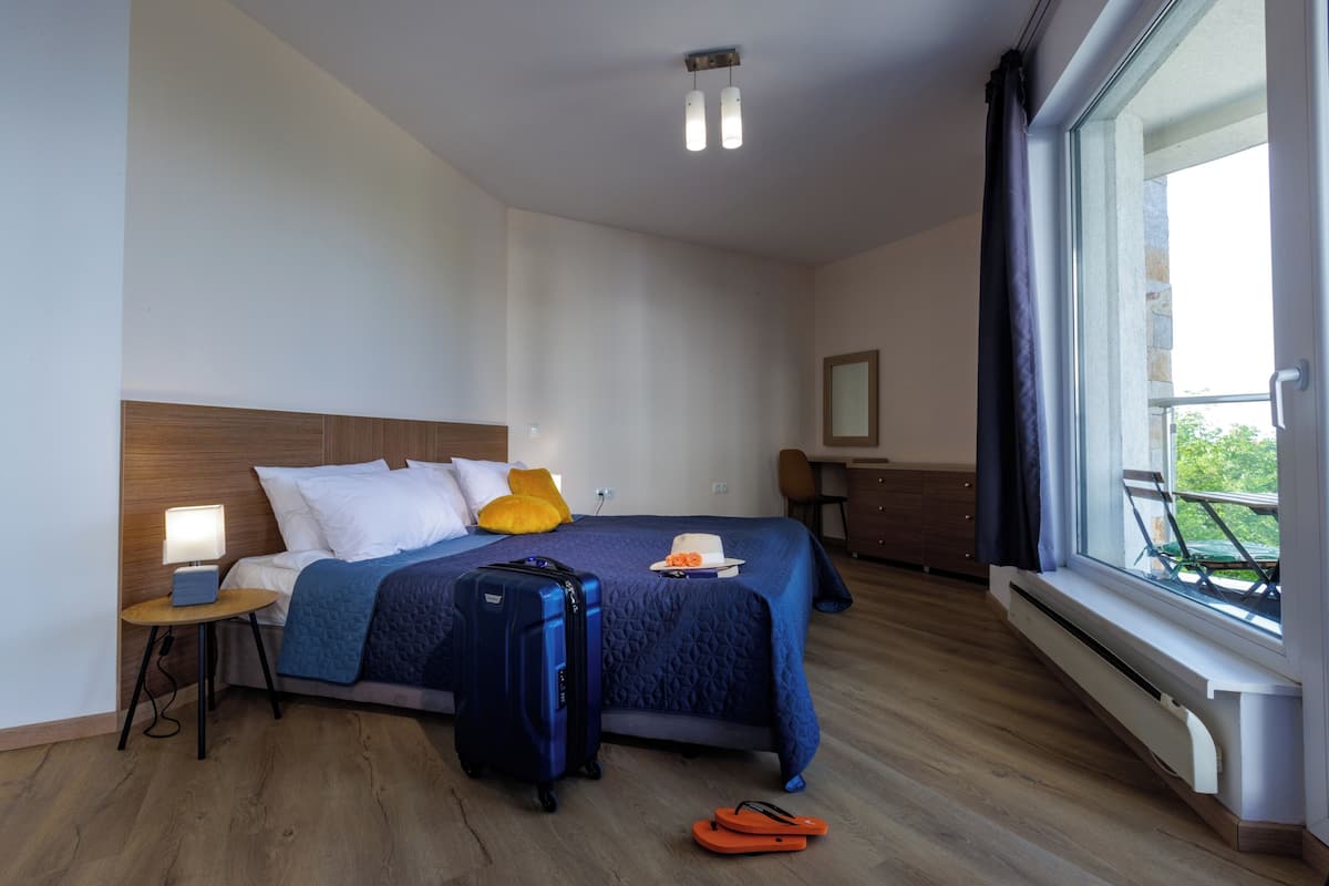 A hotel room with a bed, a suitcase, a nightstand, a lamp, a window with a view, and a chair.